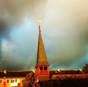 Kirketårnet belyst af solen på baggrund af blå himmel med sorte skyer og regnbue.