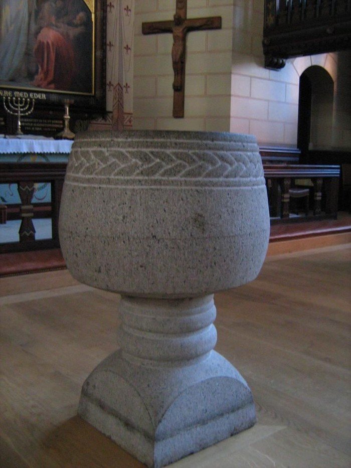 Døbefonten i grå granit med ornament langs kanten. Ligner et bægre på fod.